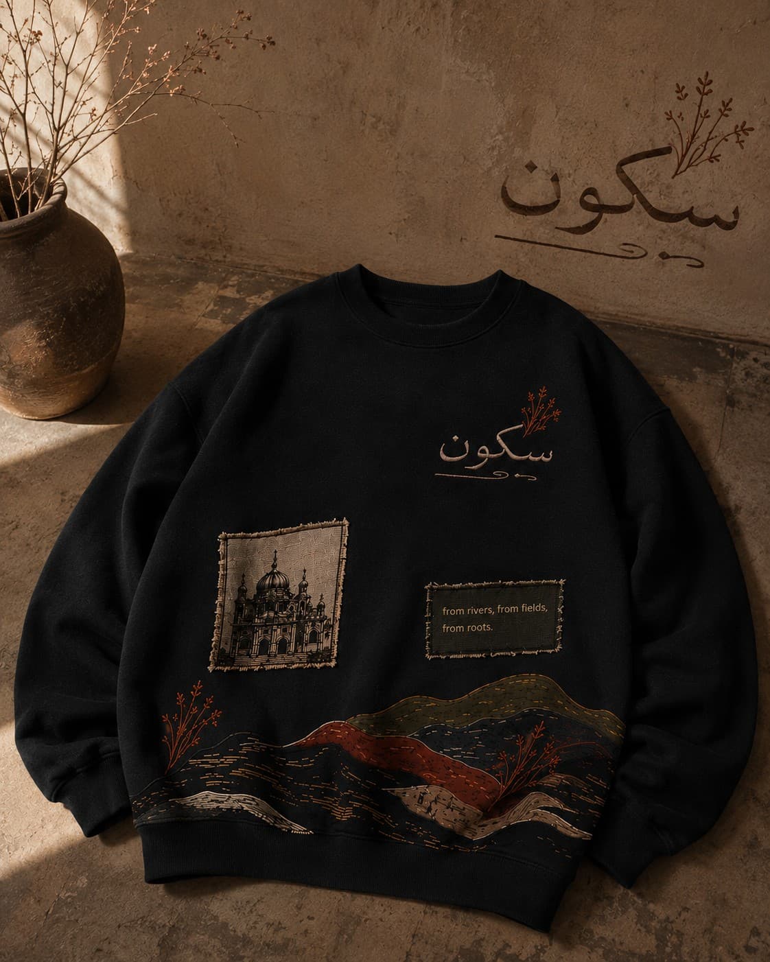 Sakoon black crewneck flat against a stone wall, embroidered سکون and mountain-river landscape motif