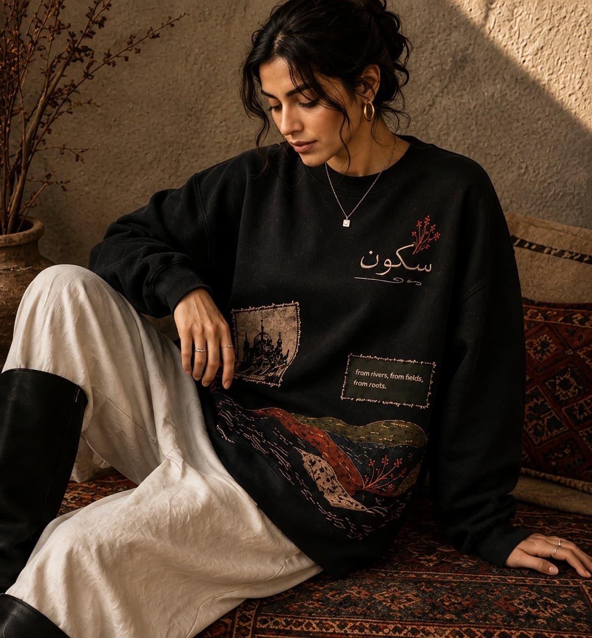 Sakoon black crewneck hung against a sunlit sandstone wall beside a woven سکون wall-hanging that reads 'from rivers, from fields, from roots'